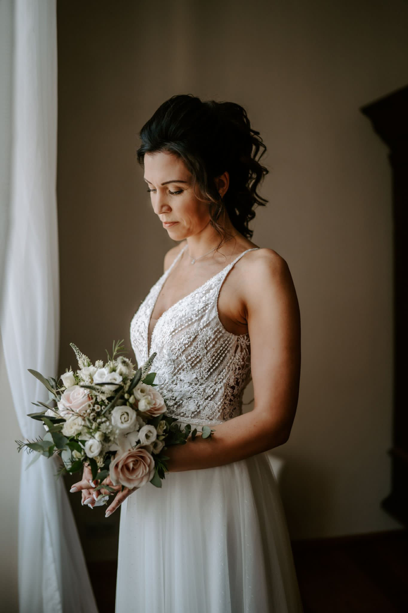 Braut mit romantischer Hochsteckfrisur, natürlichem Make-up und Blumenstrauß am Fenster